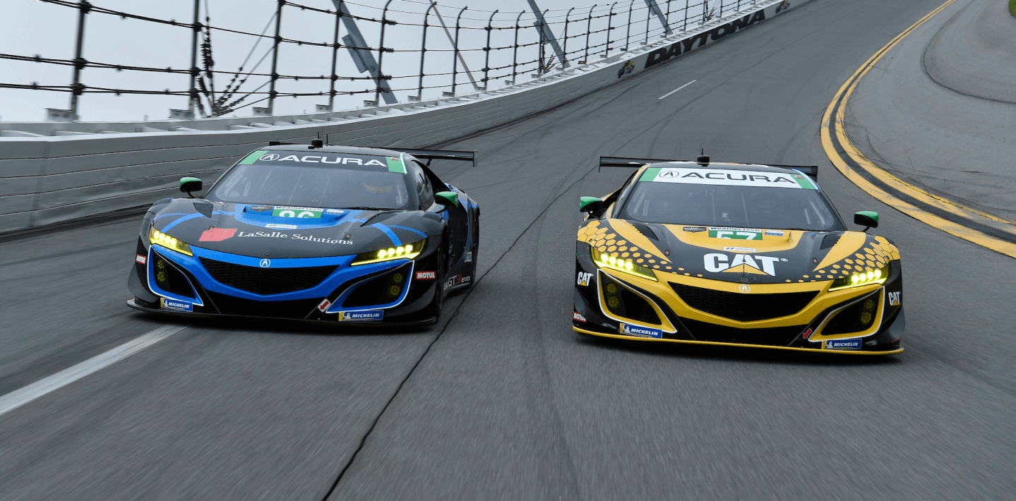 Front view of two Acura NSX GT3s, one blue and one red, both covered in sponsorship wraps, racing head-to-head on a track. Vue avant de deux Acura NSX GT3, l’une bleue et l’autre rouge, recouvertes de logos de commanditaires coursant sur un circuit de course.