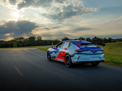 Rear, driver side angled view of an Acura TLX with red and blue racing-inspired wrap decal, driving around a track situated in an open field with trees in the distance. Vue arrière latérale du côté conducteur d’une Acura TLX recouverte d’une décalcomanie rouge et bleue roulant sur un circuit situé dans un champ avec des arbres au loin.